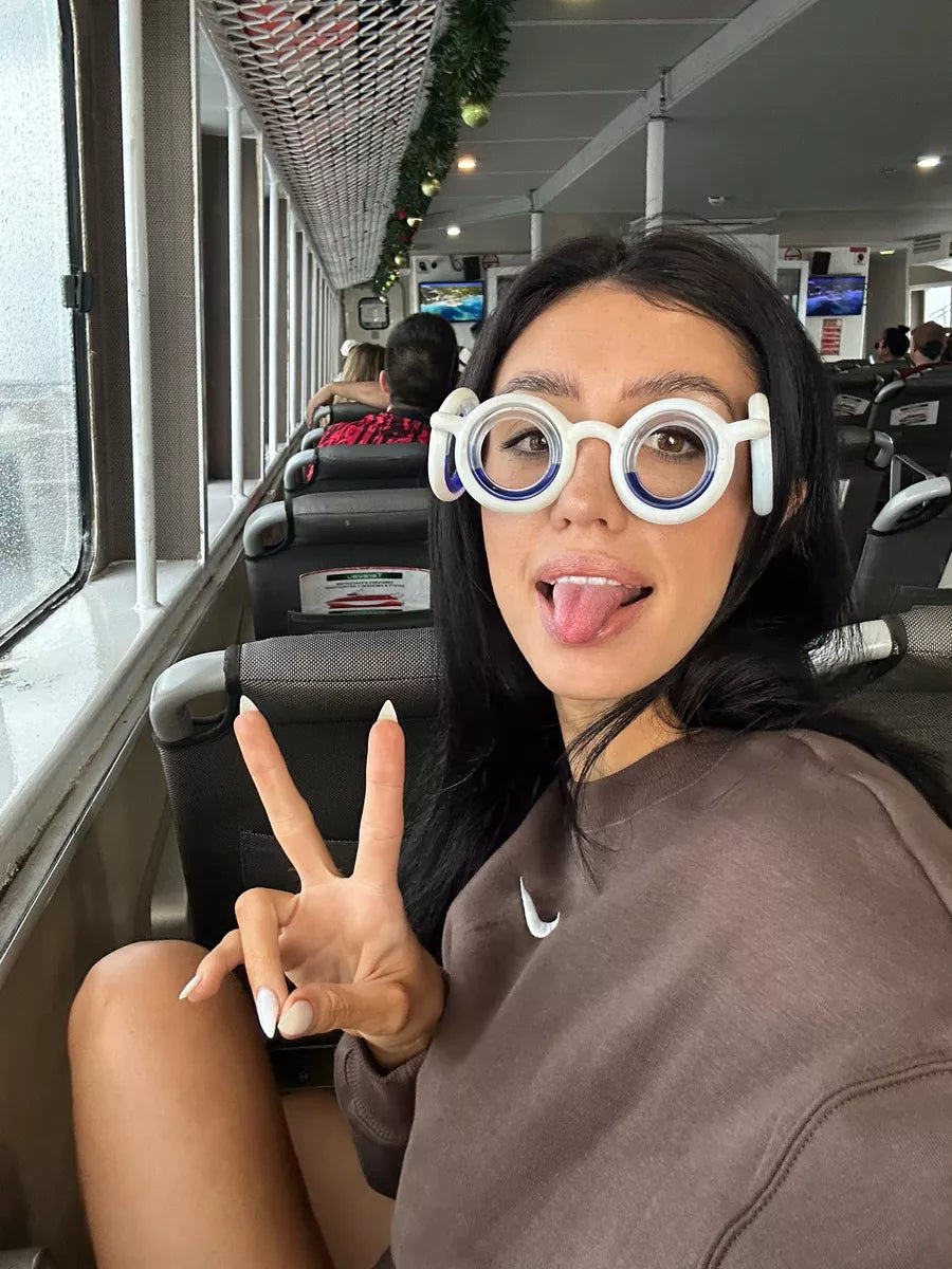 Person sitting on a bus wearing motion sickness glasses with springy eyes, making a peace sign and sticking their tongue out.
